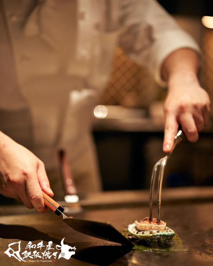 A close-up action shot of a chef in a white uniform preparing abalone on a black iron grill at a Japanese restaurant. The chef uses a spatula to place a piece of abalone onto seaweed, while another hand uses a fork to press down the topping.